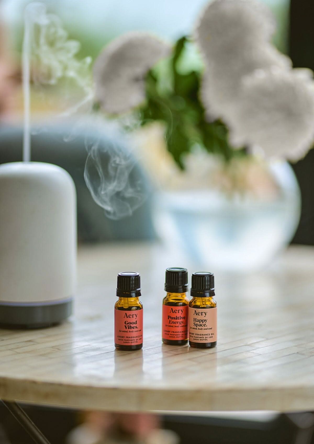 Pink Happiness Set of three fragrance oils by Aery displayed with electric diffuser and vase of flowers sat on round table